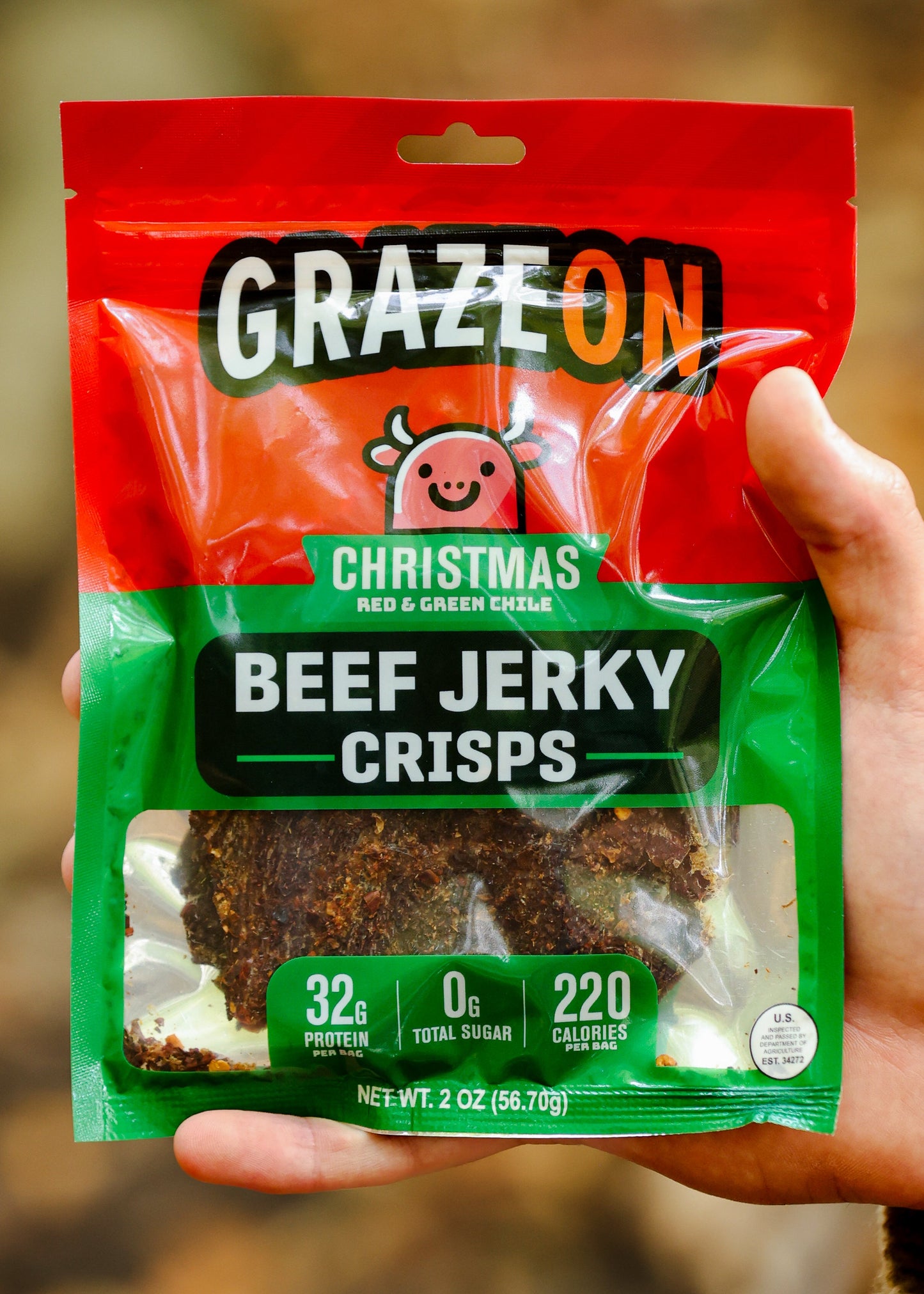 Hand holding a package of GrazeOn Christmas Beef Jerky Crisps against a blurred natural background