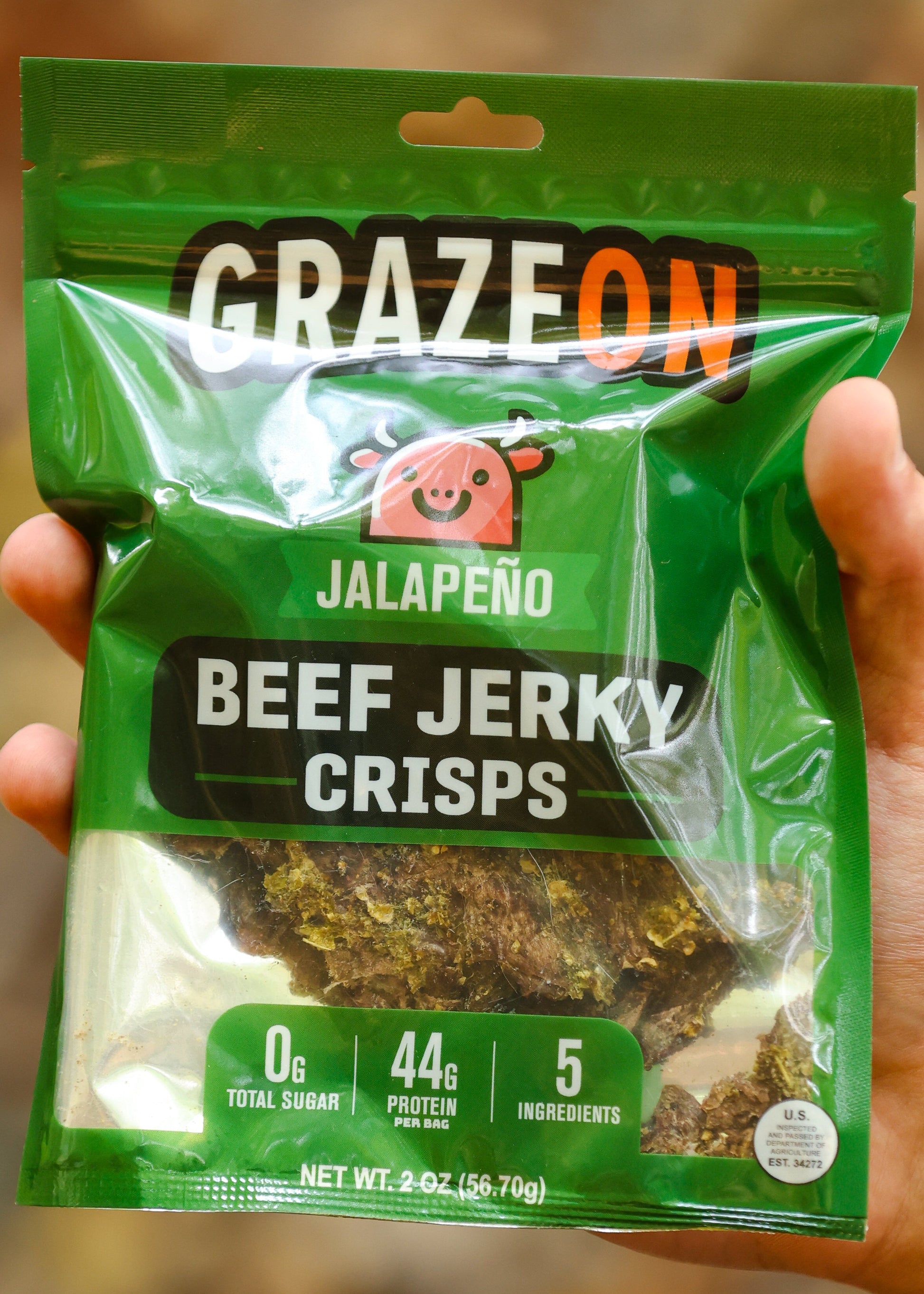 Hand holding a package of Graze On Jalapeno Beef Jerky Crisps against a blurred natural background