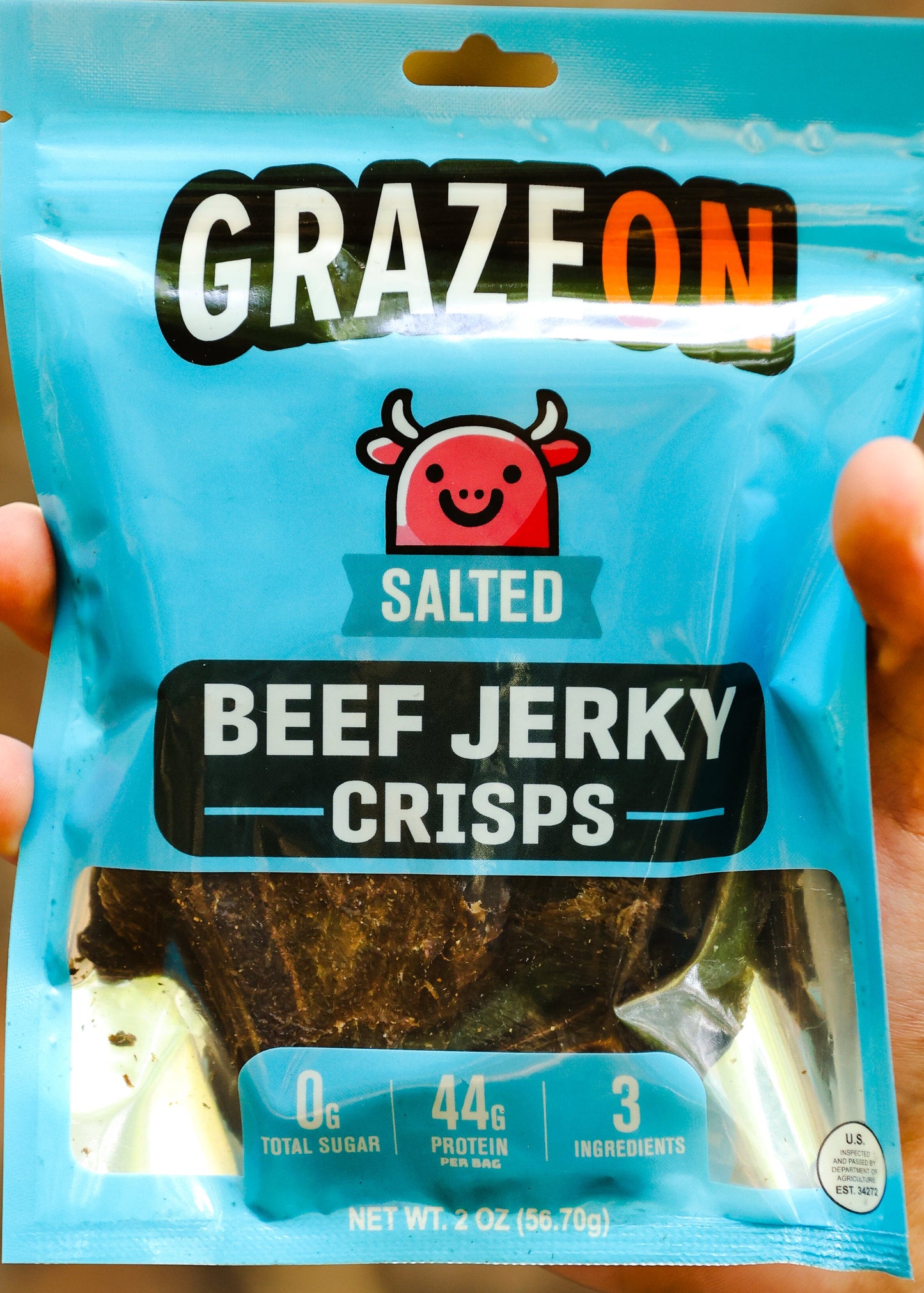 Hand holding a package of GrazeOn beef jerky crisps against a blurred natural background