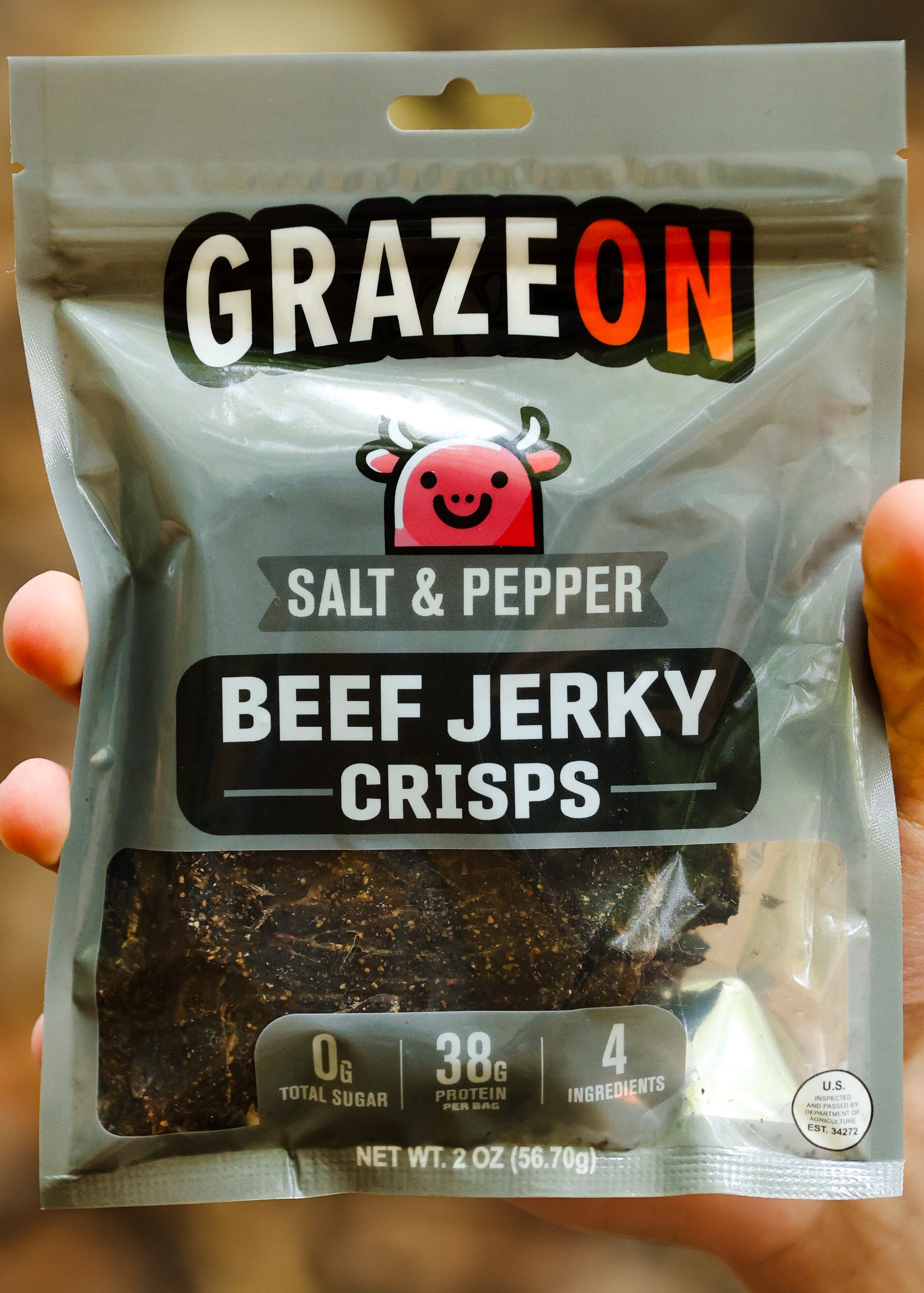 Hand holding a package of GrazeOn salt and pepper beef jerky crisps against a blurred natural background.
