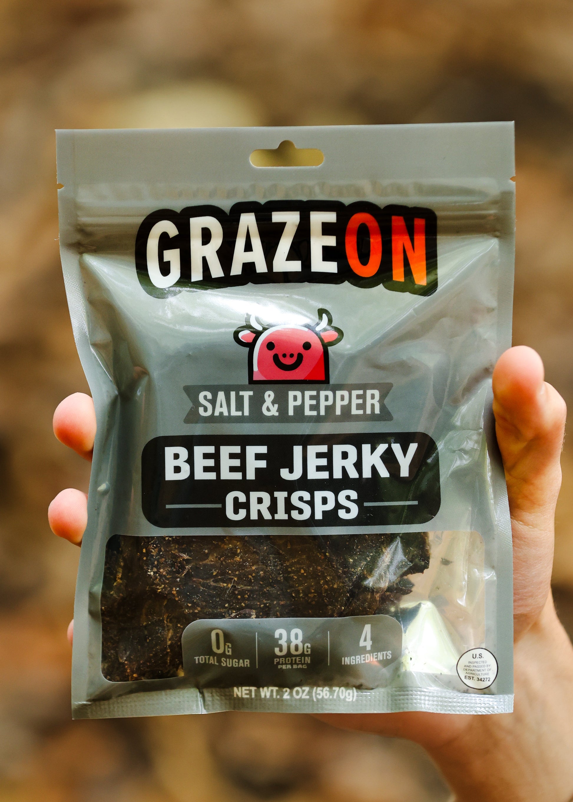 Hand holding a package of GrazeOn salt and pepper beef jerky crisps against a blurred natural background.