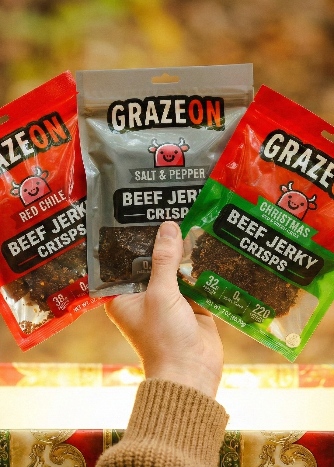Three packages of GrazeOn beef jerky held by a hand against a blurred natural background.