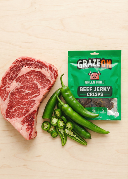 Raw beef steak, green chilies, and a package of GrazeOn Green Chile Beef Jerky Crisps on a wooden surface.