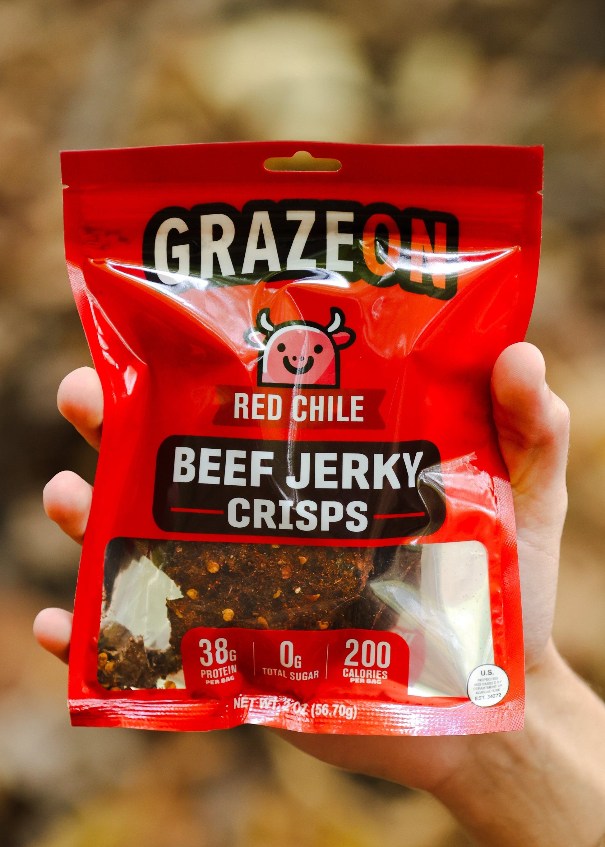 Hand holding a red package of Graze Red Chile Beef Jerky Crisps against a blurred natural background.